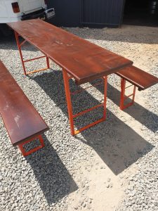 A wooden picnic table and bench with orange metal frames set on a gravel surface, with a white van partially visible in the background.